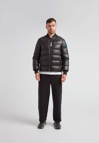 Giubbotto a vento nero con chiusura a zip, design trapuntato, polsini a coste e un accento blu sulla manica sinistra. Indossato con pantaloni neri.