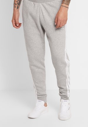Person iført lysegrå Adidas joggingbukser med hvide sidelister og hvide Adidas sneakers, stående på en ensartet lys gulv.