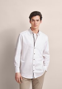 Weißes Langarmhemd mit Knopfleiste vorne, mit braunen Knöpfen und einem kontrastierenden schwarzen Akzent entlang der Knopfleiste. Glattes Baumwollmaterial.