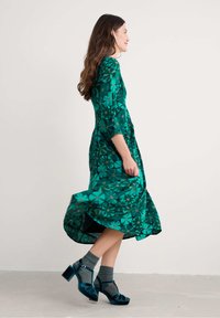 Žena prochází v zelených květinových midi šatech s 3/4 rukávy, šedými ponožkami a modrými sametovými botami na vysokém podpatku.