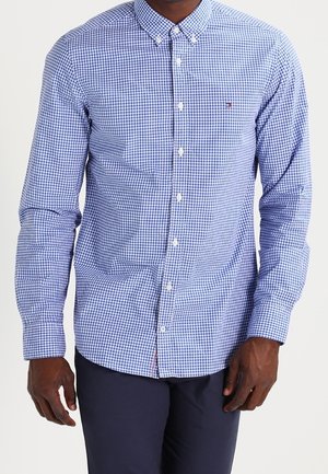 Mann trägt ein blau-weiß kariertes, langärmliges Hemd mit Knopfleiste und marineblaue Hose, steht vor einem schlichten Hintergrund.