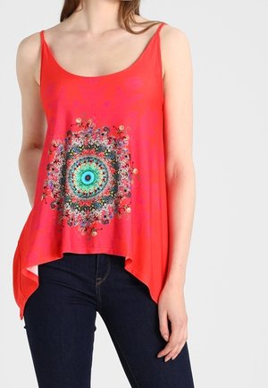 Kobieta w czerwonym topie bez rękawów z kolorowym, okrągłym wzorem mandali na środku, w połączeniu z ciemnoniebieskimi dżinsami.
