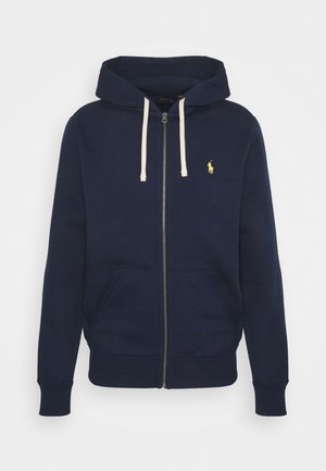 Granatowa bluza na zamek z przednimi kieszeniami, kremowymi sznureczkami, ściągaczami na rękawach oraz niewielkim żółtym haftowanym logo na lewej piersi.