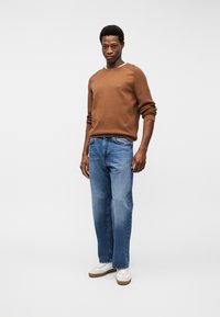 Uomo con maglione marrone, jeans blu e sneakers bianche in piedi contro uno sfondo chiaro uniforme, che guarda la macchina fotografica.