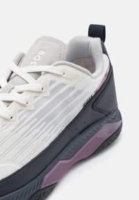 Vit och grå sneaker med svarta och lila sulaaccenter, vita snören, ventilerande mesh-sidor och vadderad krage med dragband.