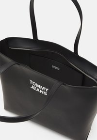 Svart tote-väska gjord av slätt material med två långa handtag, ett dragkedjefack och vit "TOMMY JEANS"-logotyp på framsidan.