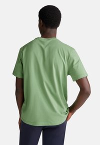 Muž s tmavou pletí oblečený v krátkém zeleném tričku a tmavých kalhotách stojí s jednou rukou na boku, otočený zády, na bílém pozadí.