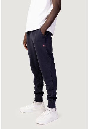 Modello maschile che indossa pantaloni da jogging blu navy con tasche laterali e piccolo logo rosso, abbinati a sneakers bianche e t-shirt bianca, in piedi con le mani nelle tasche.