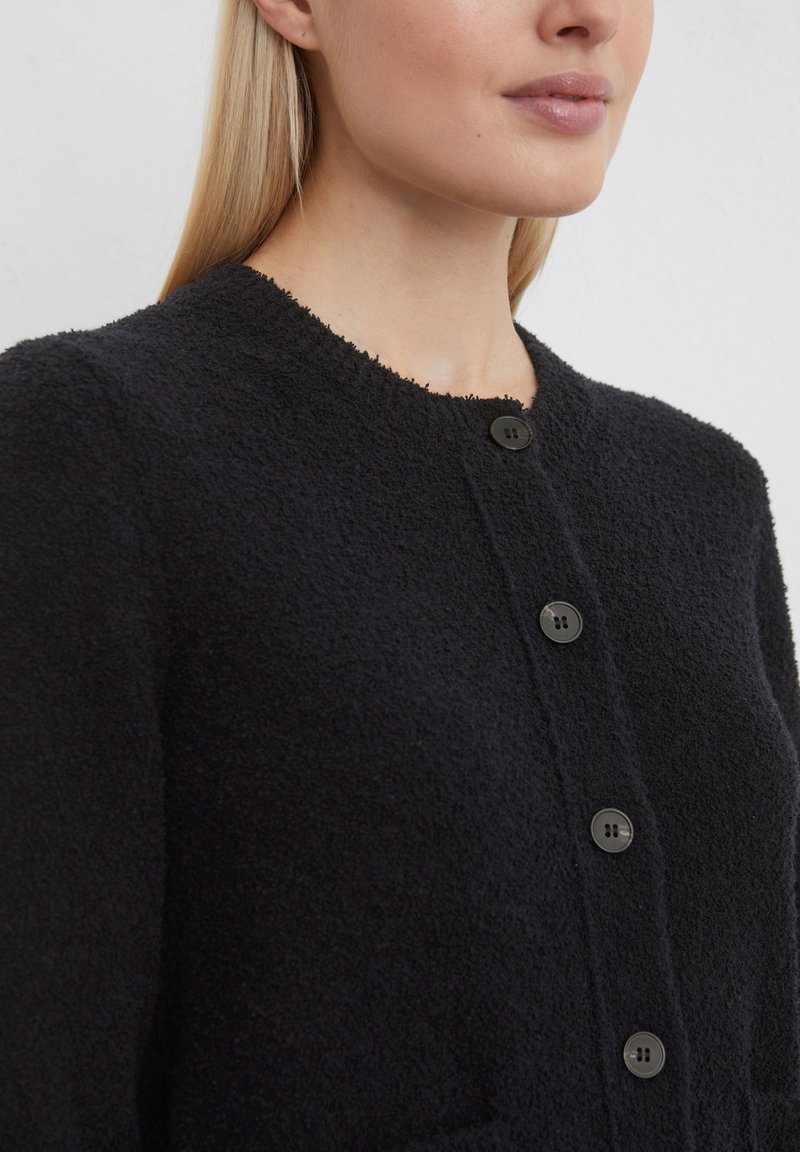 Schwarze strukturierte Strickjacke mit rundem Ausschnitt, die vorne mit fünf Knöpfen versehen ist und aus einem weichen, flauschigen Stoff besteht.