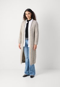 Long, open-front cardigan in light beige knit fabric, with dropped shoulders, paired with a black top, white shirt, and blue jeans.