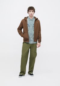 Καφέ φούτερ με κουκούλα και τσέπη μπροστά, φορεμένο πάνω από ανοιχτόχρωμο πουκάμισο από τζιν. Χακί cargo παντελόνια με πλευρικές τσέπες. Μαύρα παπούτσια με λευκές σόλες.