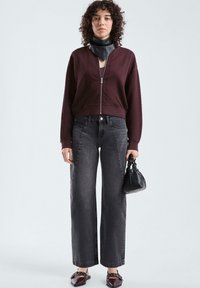 Maglione zip-up bordeaux con motivo testurizzato, jeans grigi a gamba larga, borsa nera e scarpe slip-on marroni con dettagli a strappo.