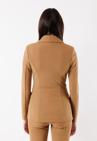 Blazer marrone su misura, caratterizzato da un design strutturato con maniche lunghe, colletto a pistagna e texture liscia. I bottoni non sono visibili nell'immagine.