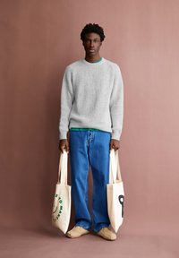 Grauer gerippter Pullover über einem grünen Hemd, kombiniert mit blauen weiten Jeans. Hält zwei beige Tragetaschen mit schwarzem und grünem Text.