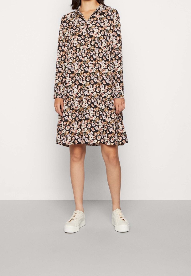 Robe noire à imprimé floral avec manches longues, col, taille cintrée et jupe longueur genou. Associée à des baskets blanches.