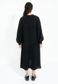 Crna haljina dugih rukava s okruglim izrezom, slobodnog kroja i dugmadima na manžetama. Glatka tekstura, dužine do polovice lista, i jednostavan dizajn na stražnjoj strani.
