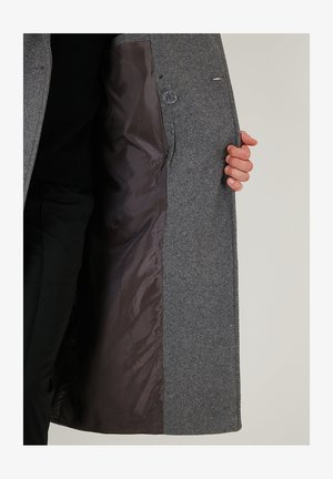 Eine Person hält einen grauen Wollmantel offen, der das glatte braune Innenfutter und einen klaren Knopf auf der Innenseite zeigt.