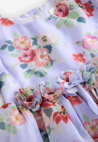 Jasnofioletowa sukienka z nadrukowanymi kwiatowymi wzorami i trójwymiarowymi materiałowymi kwiatami przyszytymi wokół linii talii.