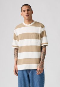 Beige and white striped t-shirt with a chest pocket, short sleeves, round neckline, made of soft fabric, worn with blue denim jeans.
