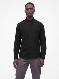 Homme portant une chemise noire à manches longues boutonnée et un pantalon gris, debout droit devant un fond blanc uni.