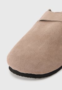 Suede slip-on sko i lys beige med avrundet tå, sort gummisåle og minimale sømdetaljer på det krysslagte overdelen.