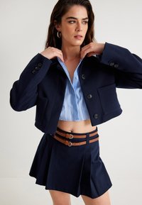 Granatowa, krótką marynarka z guzikami, w połączeniu z niebieską w paski, krótką bluzką oraz granatowymi szortami z plisami, ozdobionymi podwójnym brązowym paskiem.