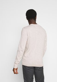 Pull beige clair avec une texture, col rond et manches longues, associé à un pantalon gris foncé. Aucun logo ou motif visible.