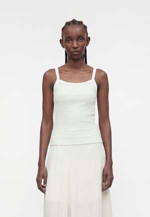 Γυναίκα με πλεγμένα μαλλιά, φοράει λευκό αμάνικο τοπ και αέρινη λευκή φούστα σε απλό φόντο.