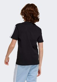 T-shirt en coton noir à manches courtes, avec des rayures blanches le long des épaules. La vue de dos montre une coupe décontractée et une texture lisse.
