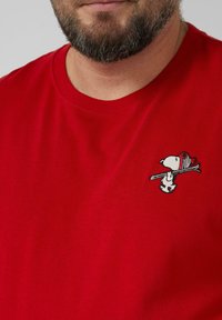 Bärtiger Mann trägt ein rotes Hemd mit einem kleinen bestickten Snoopy, der auf der linken Brust Skier trägt.