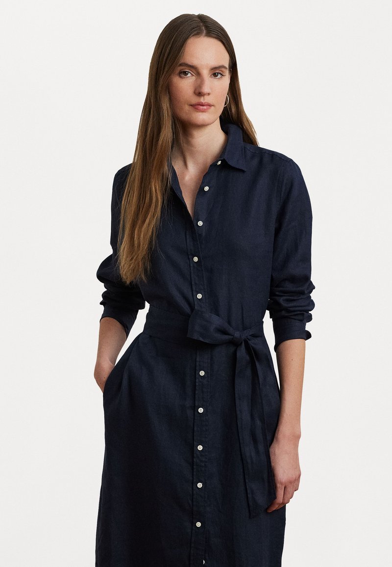 Navy blue linen shirt dress with a collar, button front, and waist tie. Features long sleeves and side pockets, showcasing a relaxed design.