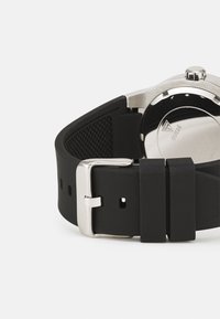 Reloj con correa de goma negra que presenta una caja y hebilla de acero inoxidable plateado. La correa tiene una superficie texturizada con múltiples agujeros de ajuste.