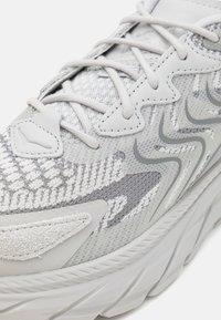 Chaussure de sport gris clair avec une tige en mesh texturé, des accents de motif subtils et une semelle rembourrée au design ondulé.