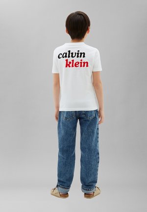 Enfant debout, de dos, portant un t-shirt blanc avec le logo « calvin klein » en noir et rouge, un jean bleu et des sandales beiges.