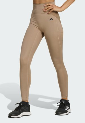 Bež legging s visokim strukom, s glatkom teksturom, detaljima šavova i crnim Adidas logom na pojasu, uparen s crnim sportskim cipelama.