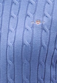 Gaiši zils džemperis ar pinuma rakstu, reljefa rakstu, ribota apmales un nelielu izšūtu logo uz krūtīm.