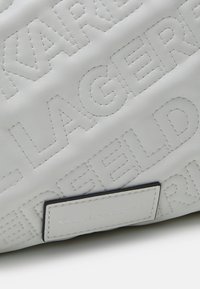 Surface en cuir blanc avec texte en relief répétitif "LAGERFELD" et une petite étiquette blanche rectangulaire avec "KARL LAGERFELD" en petits caractères.