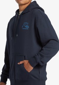Námornícky modrá mikina na zips, zmes bavlny, kapucňa na šnúrku, predná vrecko, svetlomodré logo Quiksilver na hrudi, rebrované manžety.