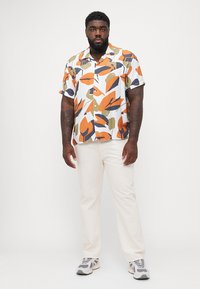Camisa de manga corta en blanco con audaces patrones de hojas en naranja, azul marino y verde oliva; combinada con pantalones de color beige claro y zapatillas grises.