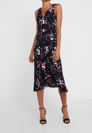 Femme portant une robe midi noire sans manches avec des motifs floraux roses et blancs et des talons noirs à bride à la cheville.