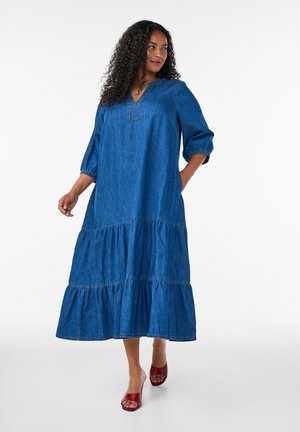 Kvinde med krøllet hår iført en løs, lagdelt blå denim midi-kjole og røde åbne hæle, stående mod en hvid baggrund.