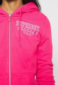 Felpa con zip rosa realizzata in tessuto morbido, dotata di tasca a marsupio, cappuccio con coulisse e testo bianco stampato sul lato sinistro del petto.