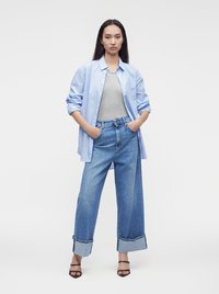 Femme debout, les mains dans les poches, portant une chemise bleu clair à boutons sur un haut gris, un jean large avec revers et des talons ouverts noirs.