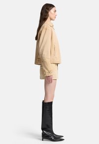 Beżowa kurtka dżinsowa o krótkim kroju, zestawiona z beżowymi szortami. Modelka nosi czarne skórzane kozaki do kolan, stoi na tle białego tła.