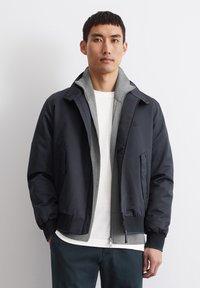 Tamsiai mėlyna bombonešio striukė su pilku vidiniu pamušalu, priekinėmis kišenėmis ir užtrauktuku. Pagaminta iš sklandaus, lengvo audinio.