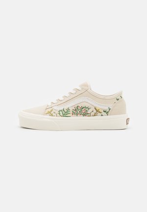 Zapatilla baja beige con franja blanca y bordado floral en los laterales, cordones blancos y suela blanca gruesa.