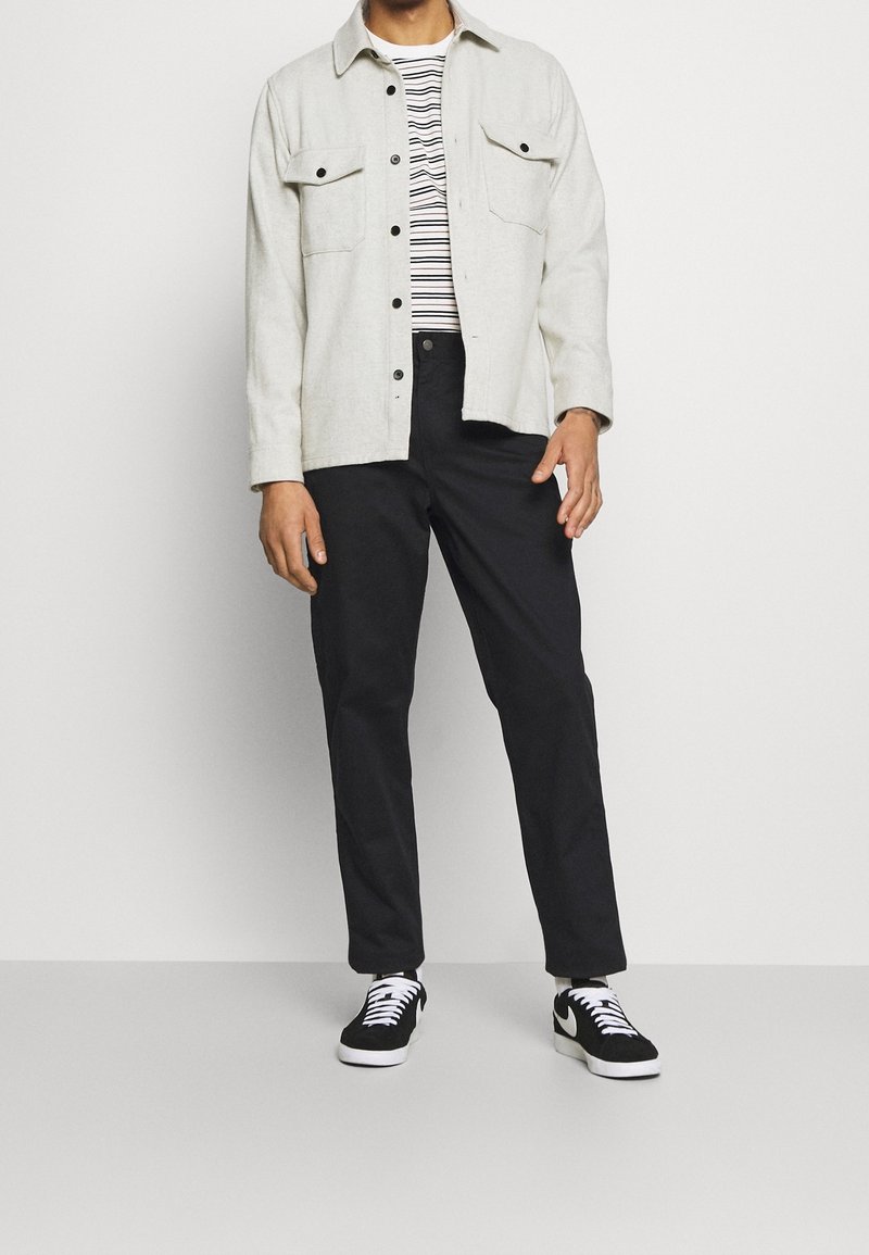 Homme portant un pantalon noir, des baskets noires et blanches, une chemise rayée et une veste beige clair boutonnée, debout devant un fond uni.