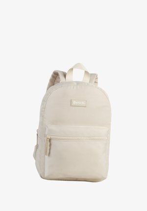Beige fabric backpack with a rounded shape, featuring a front zip pocket, padded shoulder straps, and a label with the brand "Bench."