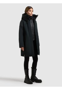 Dlouhý tmavě zelený parka s kapucí přes černou péřovou vestu, kombinovaný s černými legínami a šněrovacími bojovými botami. Hladká textura, minimalistický design.