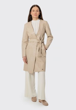 Žena oblečená v béžovém koženém kabátu s páskem, bílé plisované blůze, širokých bílých kalhotách a béžových mokasínech, stojící s jednou rukou v kapse.